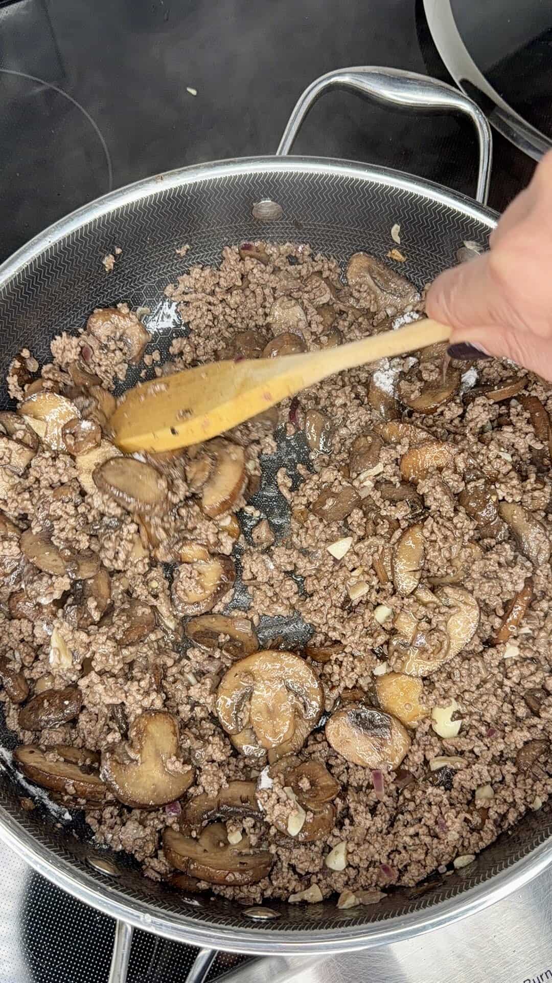 Ground beef and sliced mushrooms are being sautéed in a frying pan on a stovetop, with a hand stirring the mixture using a wooden spoon.