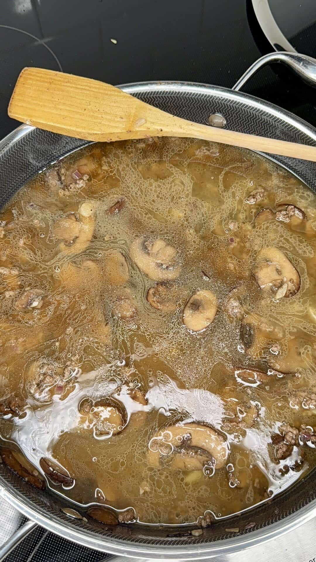 A pot of mushroom soup with visible sliced mushrooms and broth is cooking on a stovetop. A wooden spatula rests on the edge of the pot.