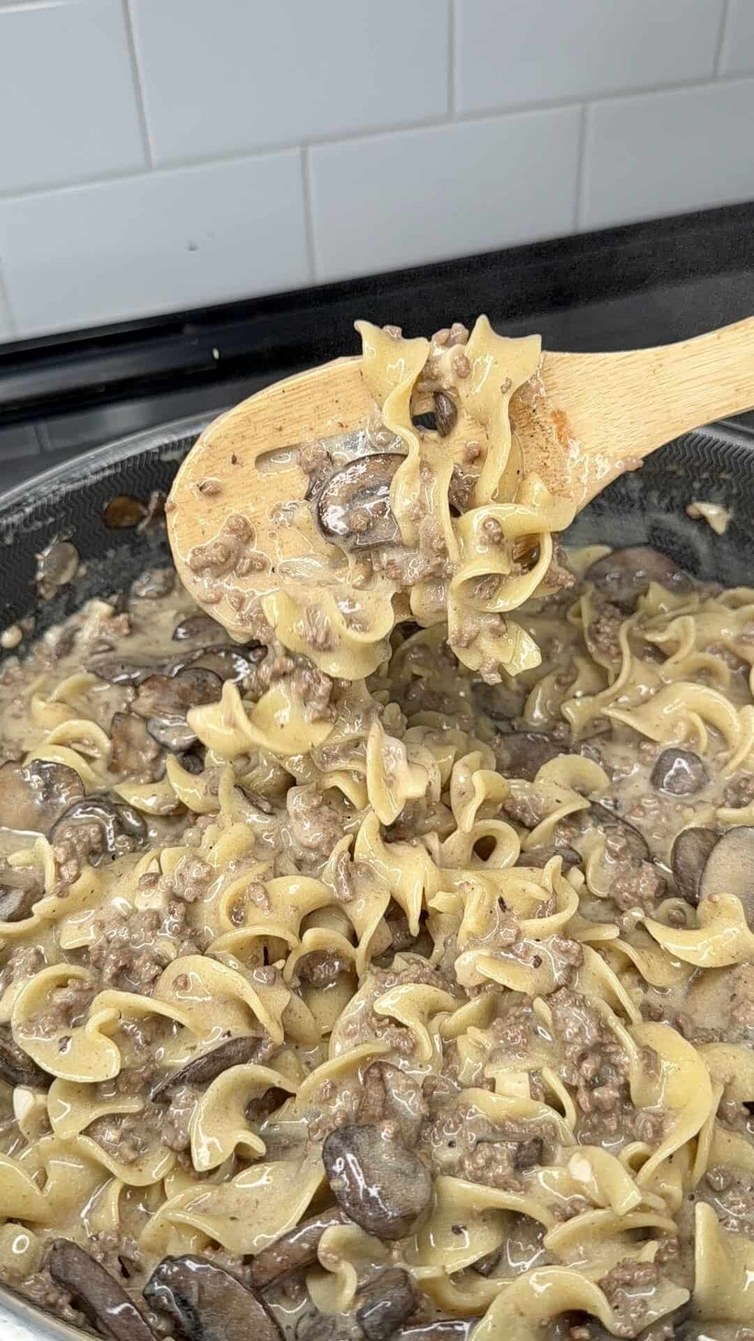 A wooden spoon lifts creamy beef and mushroom stroganoff with egg noodles from a skillet, showing the hearty mixture of ground beef, mushrooms, and sauce. White tile backsplash is visible in the background.