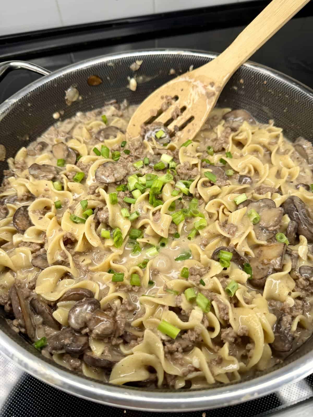 A skillet filled with creamy beef stroganoff featuring egg noodles, mushrooms, and ground beef, garnished with chopped green onions. A wooden spoon rests in the dish.