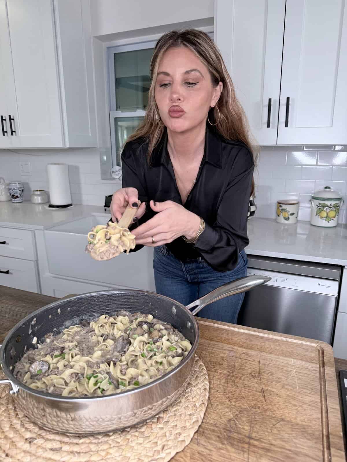 A woman with long brown hair wearing a black blouse and jeans stands in a white kitchen, serving a portion of pasta from a pan, while making a playful face with puckered lips.