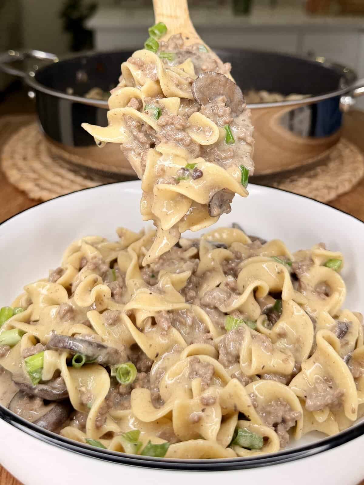 A wooden spoon holds a serving of creamy beef and mushroom stroganoff with egg noodles above a white bowl, topped with chopped green onions. A skillet of stroganoff is visible in the background.