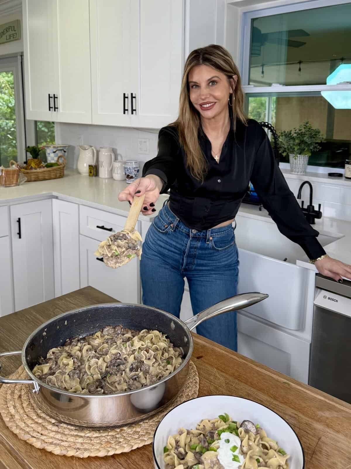 A woman in a black blouse and jeans stands in a bright kitchen, smiling as she serves a spoonful of pasta with meat and sauce from a pan on the counter into a bowl. The kitchen is clean and modern.