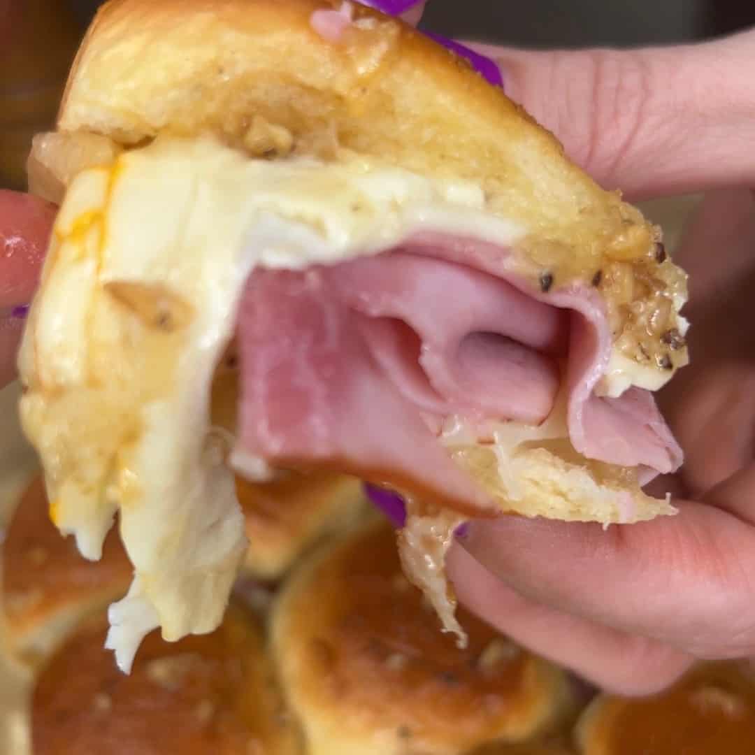 A close-up of a hand holding a ham and cheese slider with melted cheese inside a soft, golden bun. The filling is visible with layers of ham and gooey cheese.