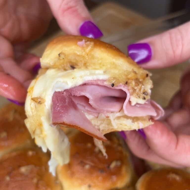 A close-up of hands with purple-painted nails holding a slider sandwich filled with ham and melted cheese on a golden bun. Other similar sliders are visible in the background.