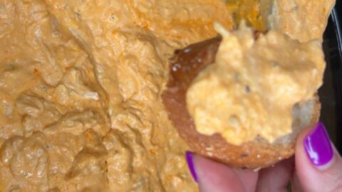 A hand with purple nail polish holds a piece of bread topped with creamy orange dip, above a bowl filled with the same dip.