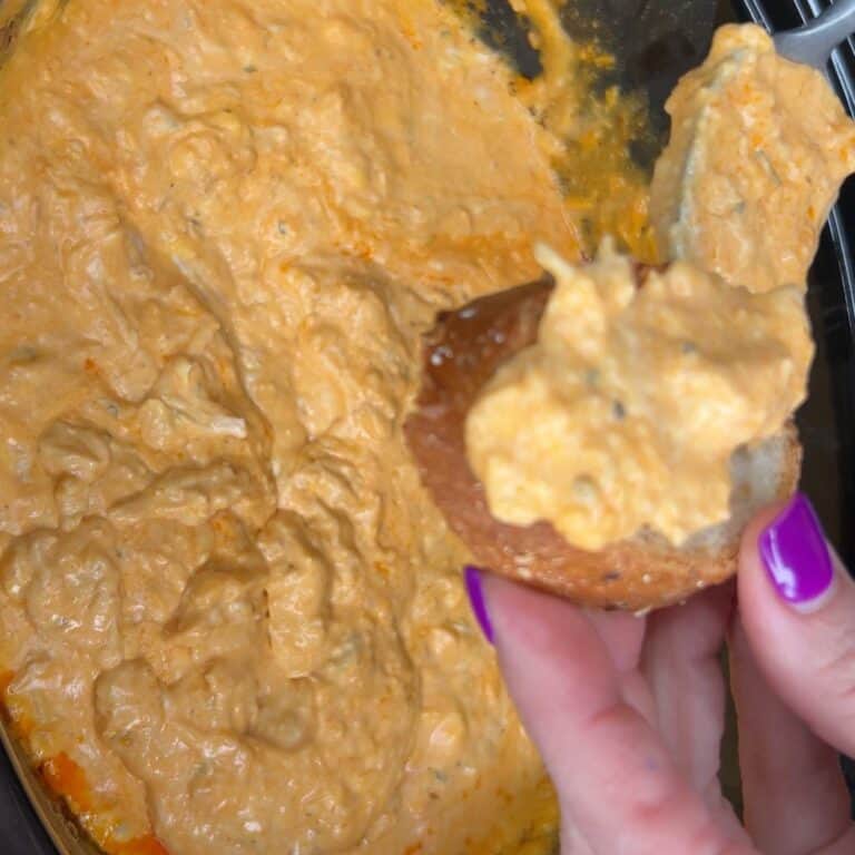 A hand with purple nail polish holds a piece of bread topped with creamy orange dip, above a bowl filled with the same dip.