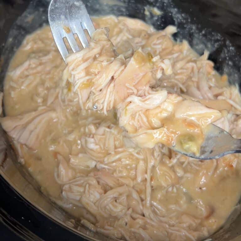 Close-up of shredded chicken being pulled apart with two forks in a thick, creamy sauce, likely inside a pot or slow cooker.