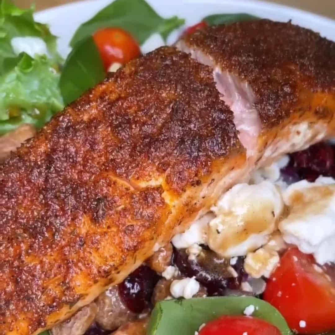 A close-up of a seasoned, blackened salmon fillet served on a salad with cherry tomatoes, leafy greens, crumbled feta cheese, and mushrooms.