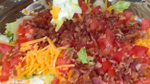 A hand with pink nail polish holds a chip scooping layered dip topped with chopped lettuce, tomato, shredded cheddar cheese, and bacon from a glass dish.