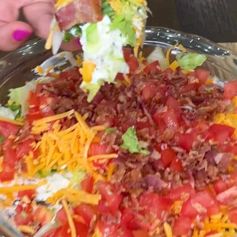 A hand with pink nail polish holds a chip scooping layered dip topped with chopped lettuce, tomato, shredded cheddar cheese, and bacon from a glass dish.