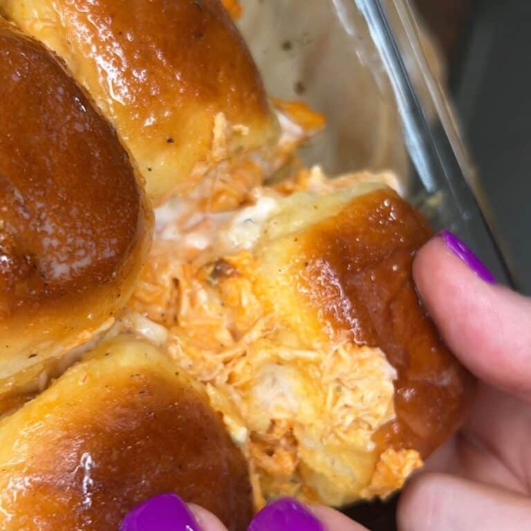 A close-up of hands with purple nail polish pulling apart a cheesy buffalo chicken slider with golden, glossy buns.