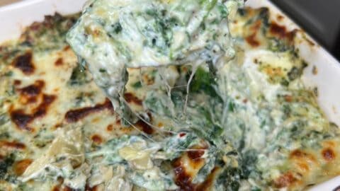 A close-up of a spoon lifting a serving of creamy, cheesy spinach and artichoke dip from a baked dish, with melted cheese stretching and a golden, bubbly top visible.