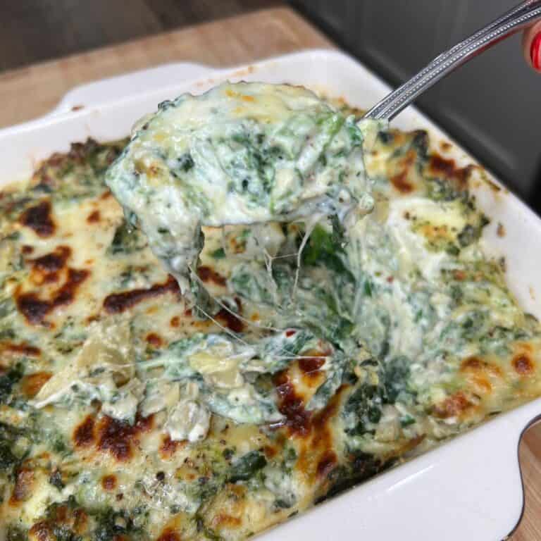 A close-up of a spoon lifting a serving of creamy, cheesy spinach and artichoke dip from a baked dish, with melted cheese stretching and a golden, bubbly top visible.