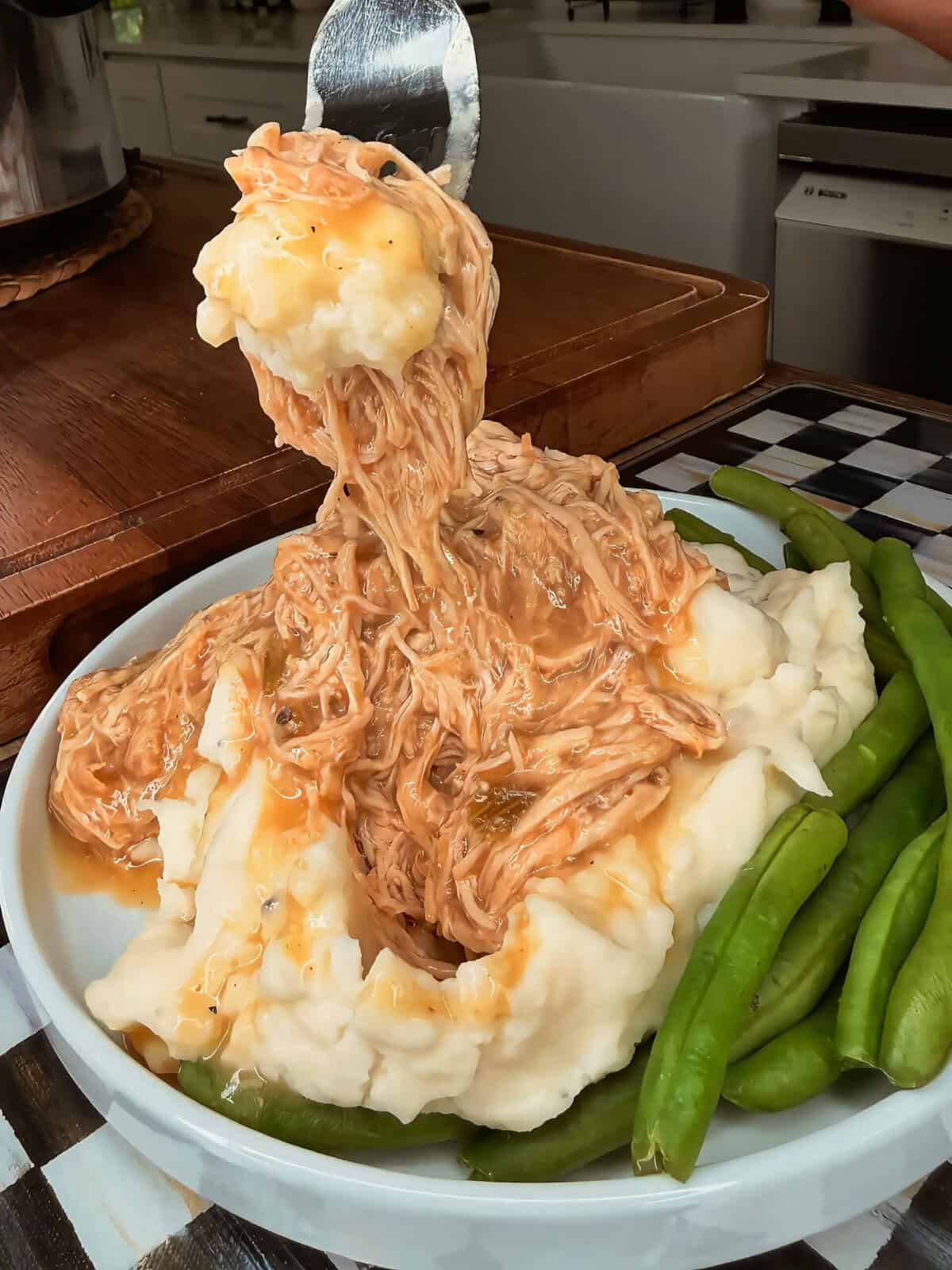 A plate of mashed potatoes topped with shredded chicken and gravy, surrounded by fresh green beans. A serving spoon lifts some chicken and potatoes from the plate.