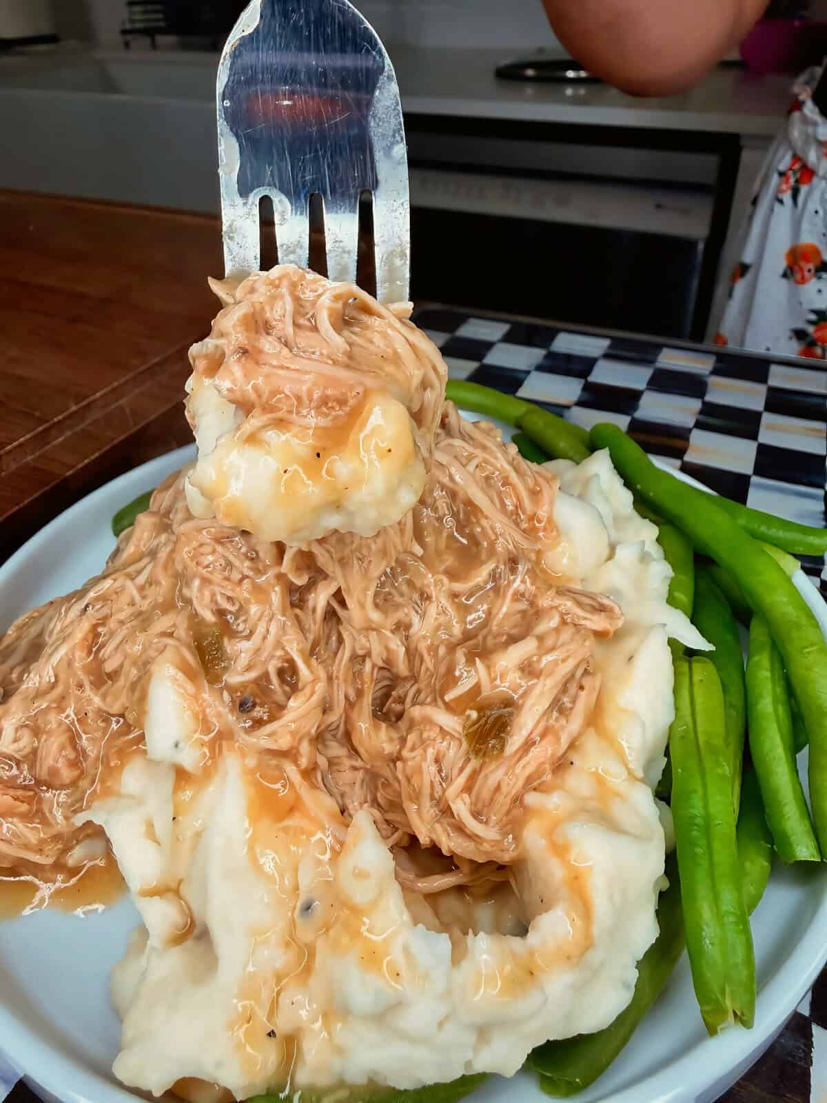 A serving of mashed potatoes topped with shredded chicken and gravy, with green beans on the side, held up by a spatula over a white plate.