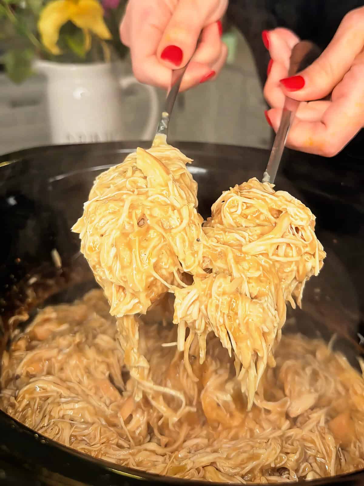 Two hands with red-painted nails lift creamy shredded chicken and noodles from a black crockpot using two forks. Flowers in a white vase are blurred in the background.