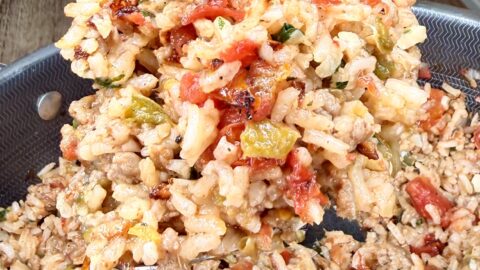 A close-up of a spoonful of cooked rice mixed with ground meat, tomatoes, and diced bell peppers being lifted from a pan. The background shows a white container on a wooden table.