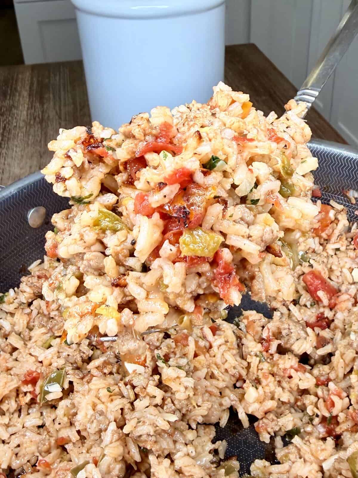 A close-up of a spoonful of cooked rice mixed with ground meat, tomatoes, and diced bell peppers being lifted from a pan. The background shows a white container on a wooden table.