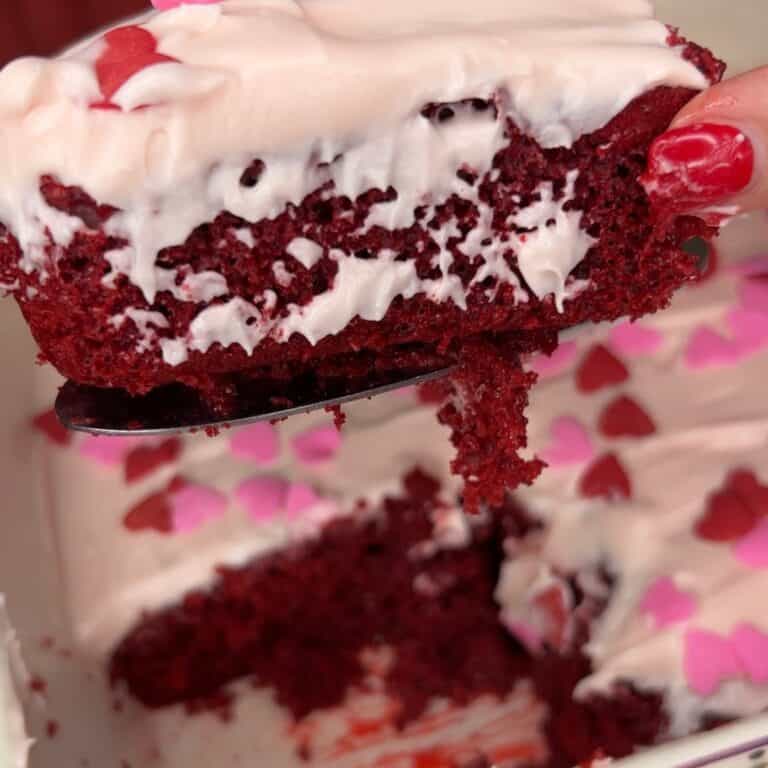 A close-up of a slice of red velvet cake with white cream cheese frosting and pink heart-shaped sprinkles, being lifted from a cake. A hand with red nail polish holds the serving utensil.