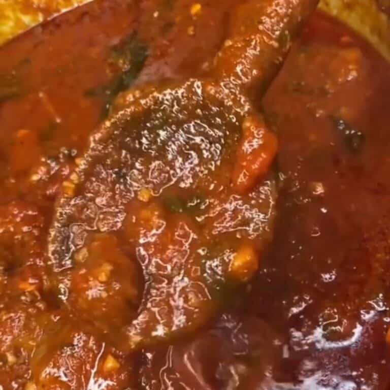 A close-up of a wooden spoon stirring a thick, chunky tomato sauce with visible herbs and pieces of vegetables. The sauce looks rich and glossy.