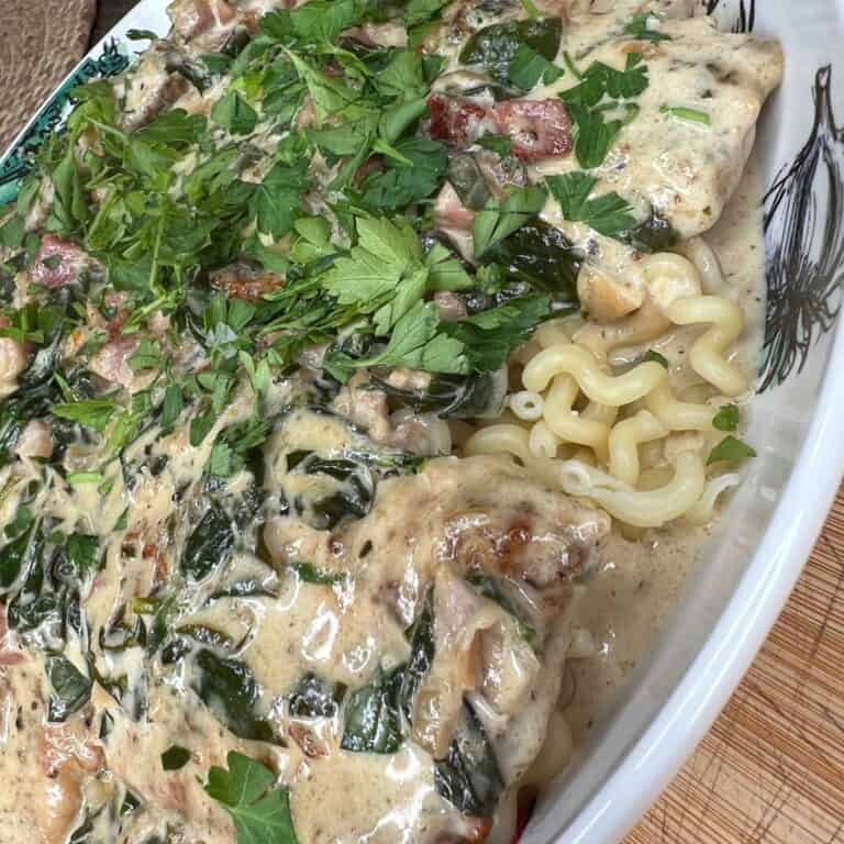 A close-up of creamy pasta topped with chopped herbs, spinach, diced tomatoes, and a rich white sauce, served in a white dish. The pasta is curly and the sauce covers the ingredients generously.