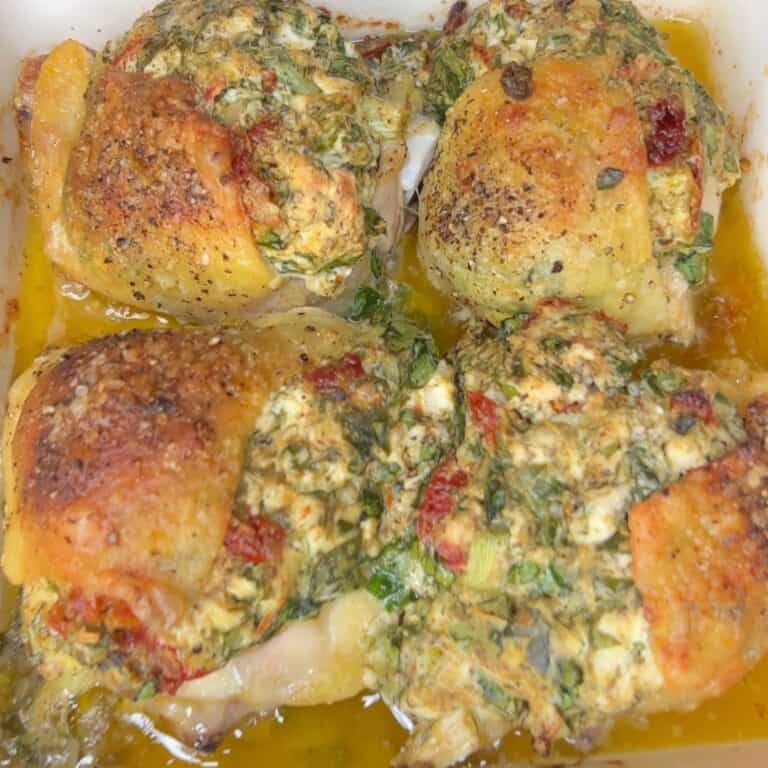 Four baked chicken thighs topped with a mixture of herbs, cheese, and seasonings, sitting in a pan with golden juices and a crispy, browned skin.