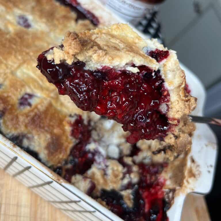 A close-up of a spoon holding a serving of berry cobbler with a golden, flaky crust and gooey mixed berry filling, above a white baking dish filled with more cobbler.