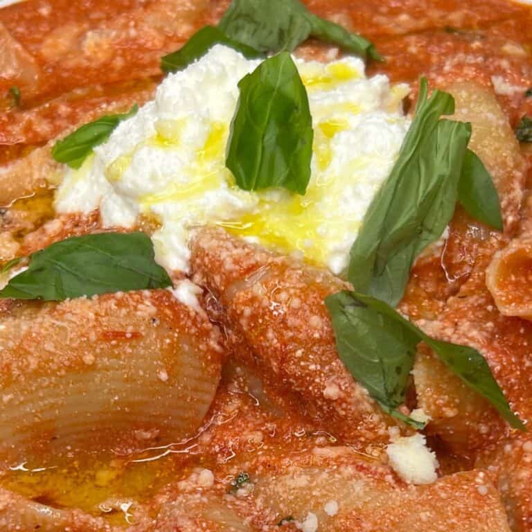 Close-up of rigatoni pasta with tomato sauce, topped with a dollop of ricotta cheese, drizzled with olive oil, and garnished with fresh basil leaves.