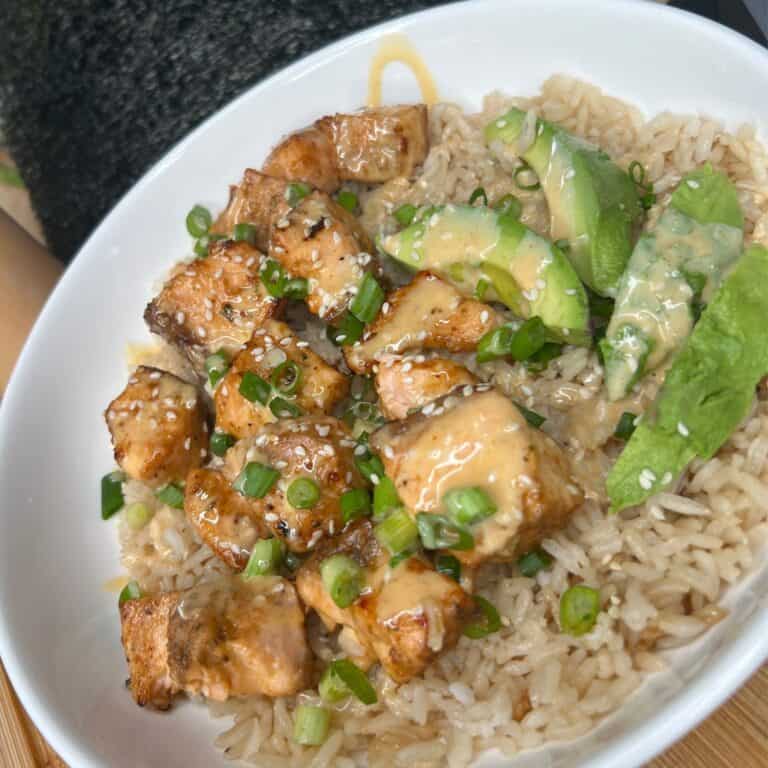 A white bowl filled with brown rice, grilled salmon chunks, avocado slices, chopped green onions, and a creamy sauce drizzled on top.