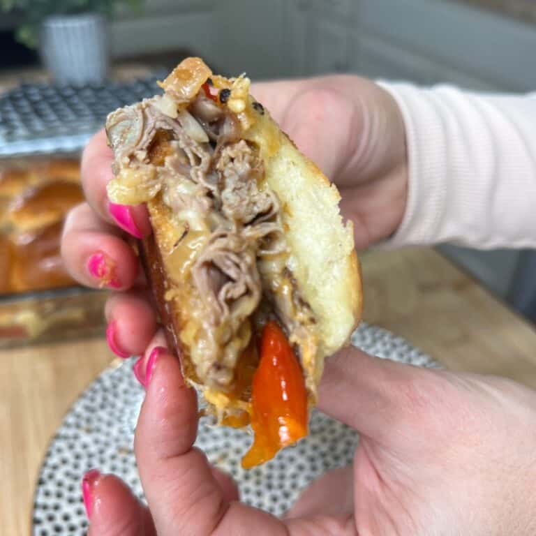 A close-up of a hand holding a sandwich filled with shredded meat, melted cheese, and red bell pepper; the sandwich is slightly squeezed, showing its savory contents.