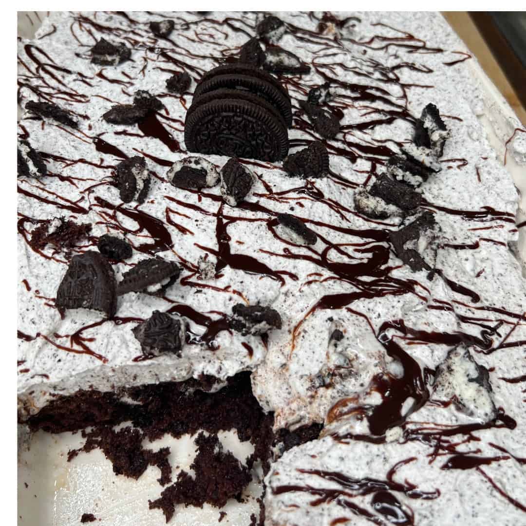 A chocolate cake topped with cookies and cream frosting, crumbled chocolate sandwich cookies, and a drizzle of chocolate syrup. A piece has been cut out from the corner of the cake.