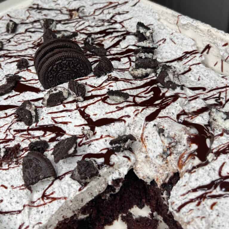 A close-up of a cookies and cream dessert topped with whipped cream, crushed chocolate sandwich cookies, whole cookies, and drizzled with chocolate syrup. A piece has been cut out, revealing a dark chocolate cake layer underneath.