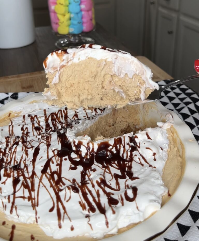 An EASY Biscoff Peanut Butter Pie with a creamy, light brown filling and whipped topping drizzled with chocolate syrup is being served; a spatula lifts a slice onto a patterned plate, with colorful marshmallow treats in the background.