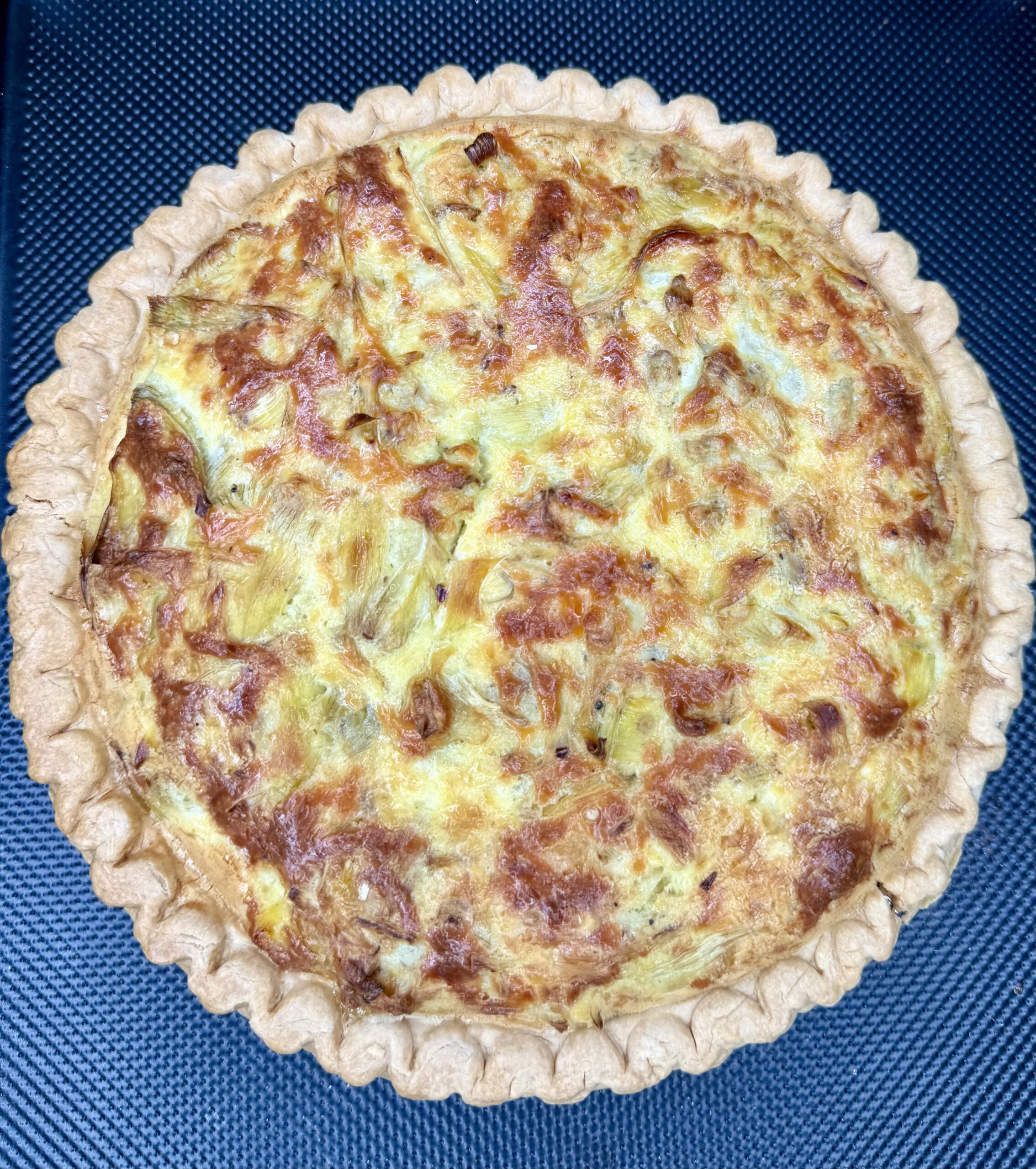 Grandma Jo's Artichoke Quiche, with its golden, flaky crust and cheesy, browned filling, sits invitingly on a dark textured surface.