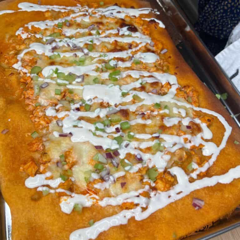Rectangular golden-brown flatbread topped with melted cheese, diced chicken, chopped red onions, green onions, and drizzled with white sauce, served on a metal tray.