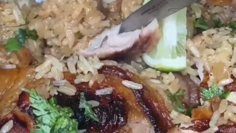 A close-up of a fork and knife cutting into grilled chicken served over seasoned rice, garnished with fresh cilantro and lime slices. A hand with red nail polish holds the knife.