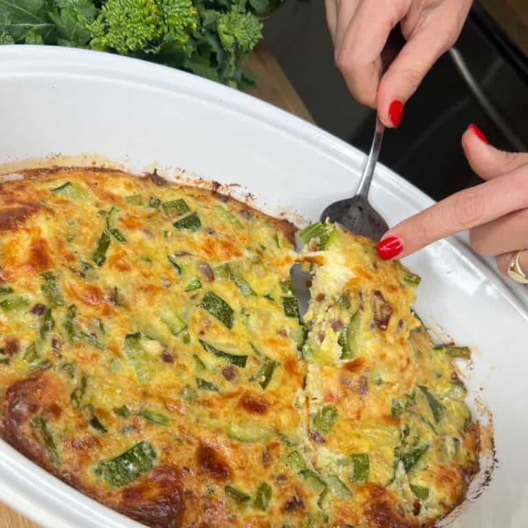 A person with red nail polish scoops out a serving of what could be the BEST Vegetable Frittata casserole, likely zucchini, from a white dish. Fresh greens are visible in the background on a wooden countertop.