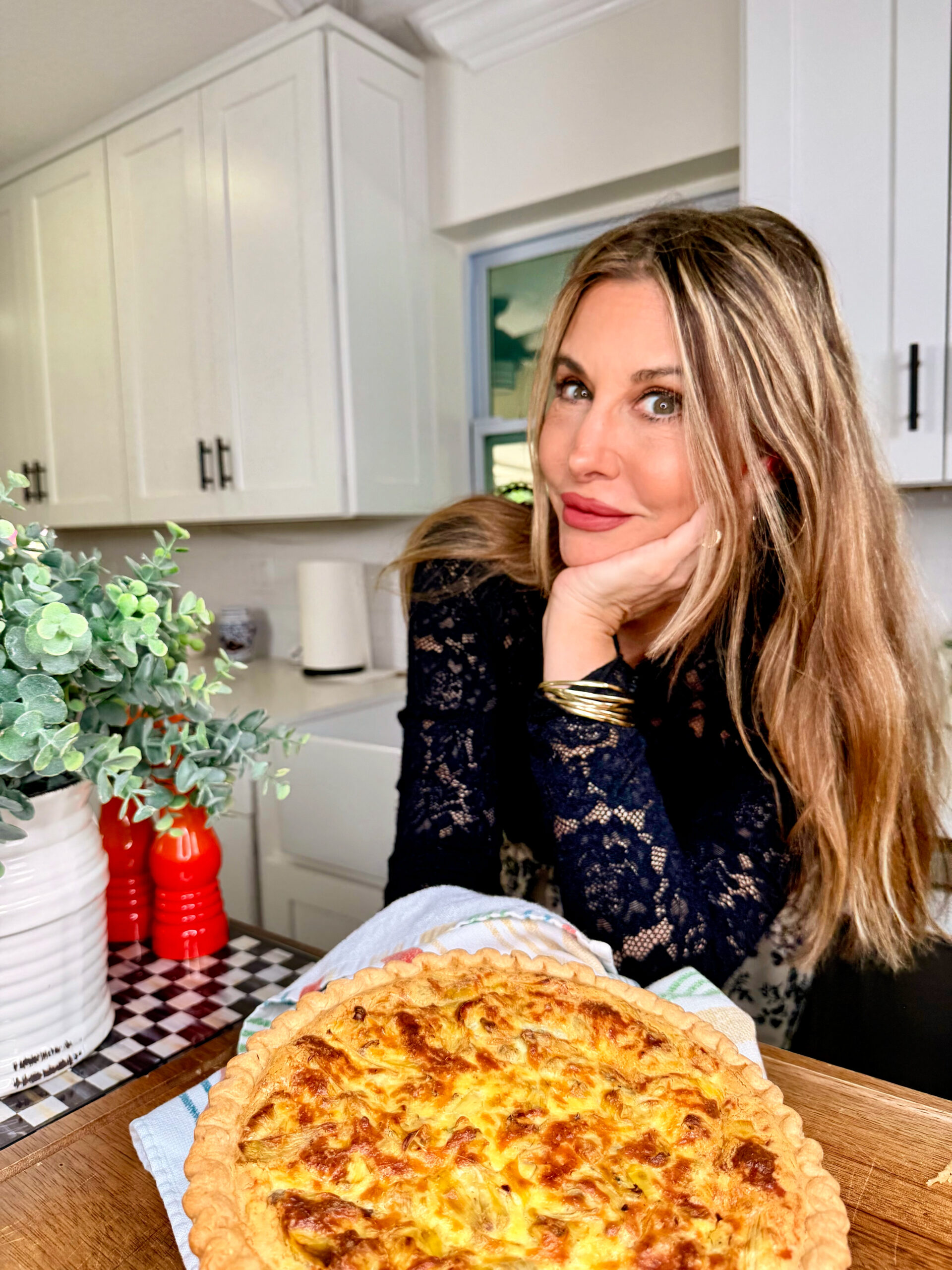 A woman with long blonde hair, wearing a dark lace top, smiles and rests her chin on her hand in a kitchen next to Grandma Jo's Artichoke Quiche on a wooden board. Greenery and kitchen items are visible in the background.