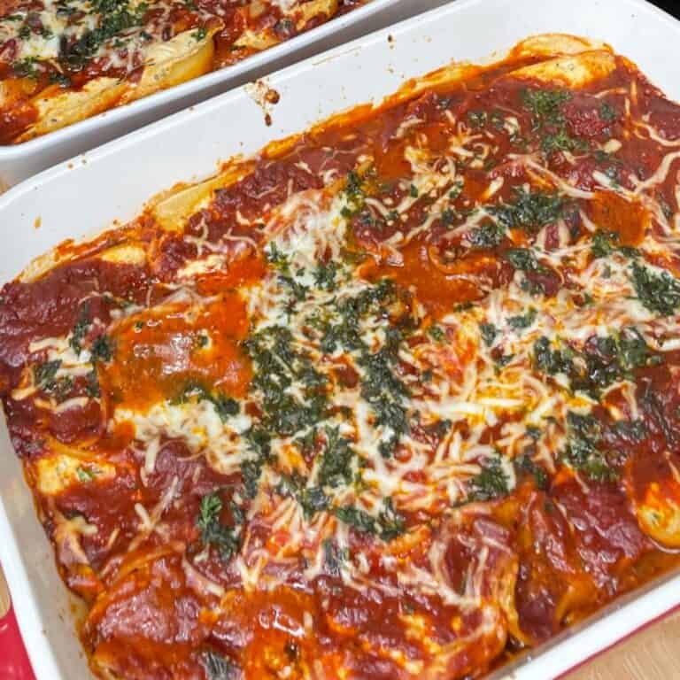A close-up of a baked lasagna in a white dish, topped with melted cheese, tomato sauce, and sprinkled herbs. The edges are slightly browned, with another similar dish visible in the background.