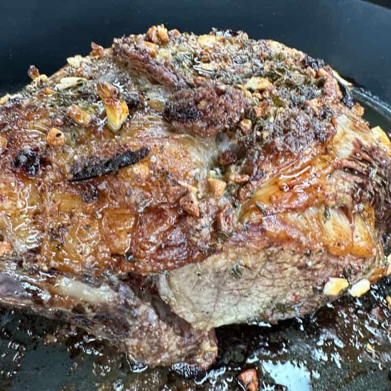 A close-up of a roasted beef roast with a browned, crispy exterior, topped with chopped garlic and herbs, sitting in a pan with juices.
