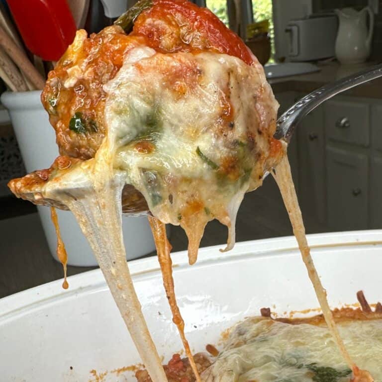A spoon lifts a cheesy, saucy meatball from a white dish, with melted cheese stretching down. The background shows a kitchen with utensils in a container.