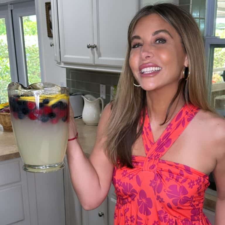 A smiling woman in a pink floral dress holds a large pitcher of lemonade with ice, lemon slices, blueberries, and blackberries in a bright kitchen.
