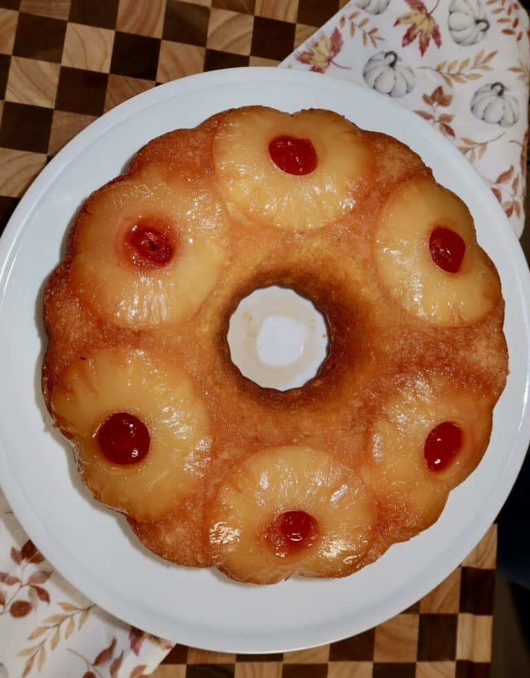 A pineapple upside-down bundt cake topped with pineapple rings and maraschino cherries sits on a white plate over a patterned wooden surface and a pumpkin-printed cloth.