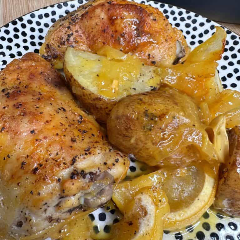 Roasted chicken pieces with crispy skin served alongside baked potato wedges and lemon slices, all covered in a glossy, seasoned sauce on a black and white dotted plate.