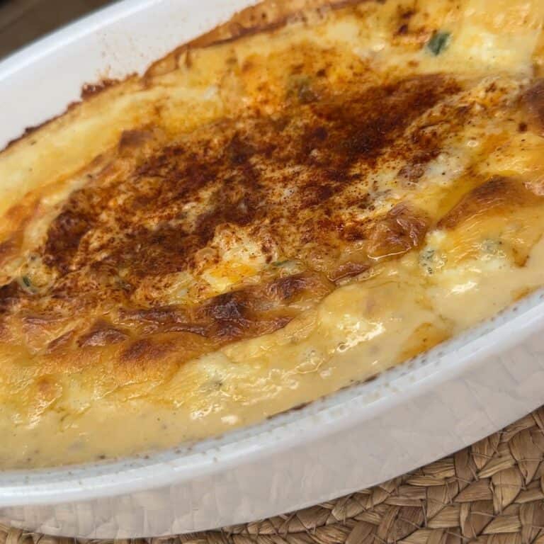 A close-up of a baked casserole in a white dish, featuring a golden-brown, bubbly, cheesy crust. The dish rests on a woven placemat.