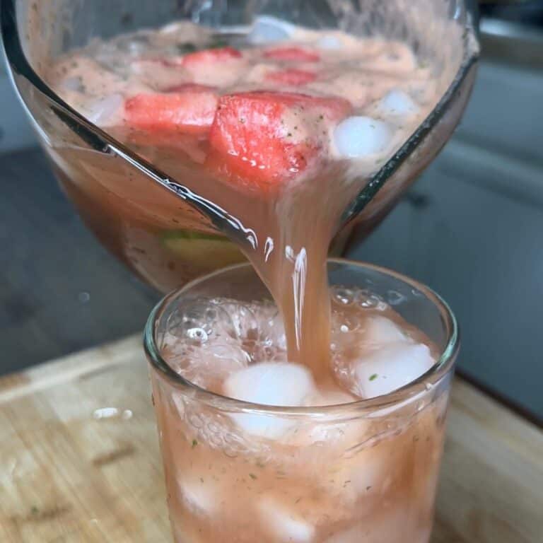 A pitcher pours a pinkish fruit drink with ice and watermelon chunks into a glass filled with ice, set on a wooden surface.