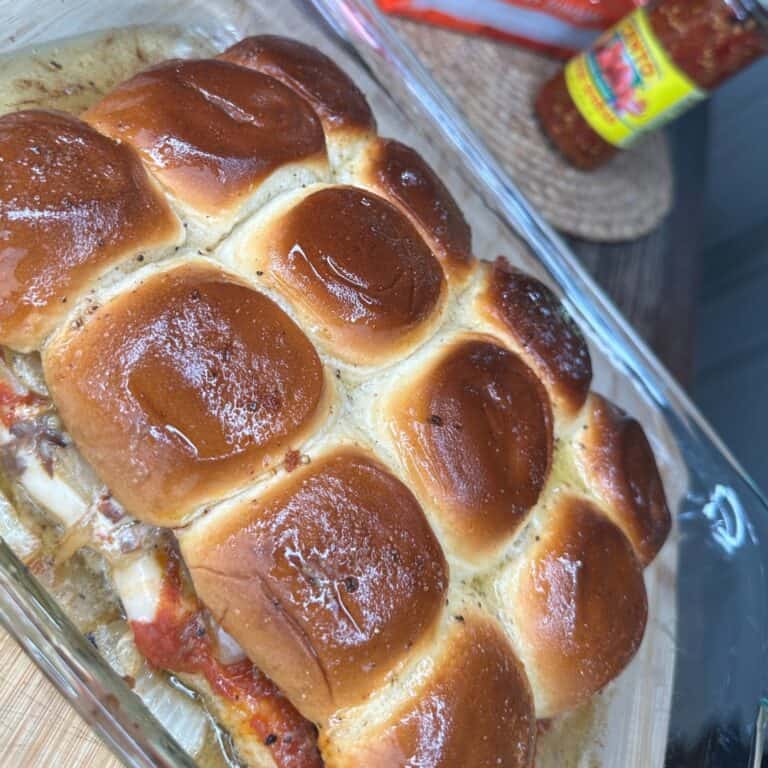A glass baking dish filled with golden-brown baked slider buns, with melted cheese and sauce visible at the edges, next to a bottle of Giardiniera relish on a table.