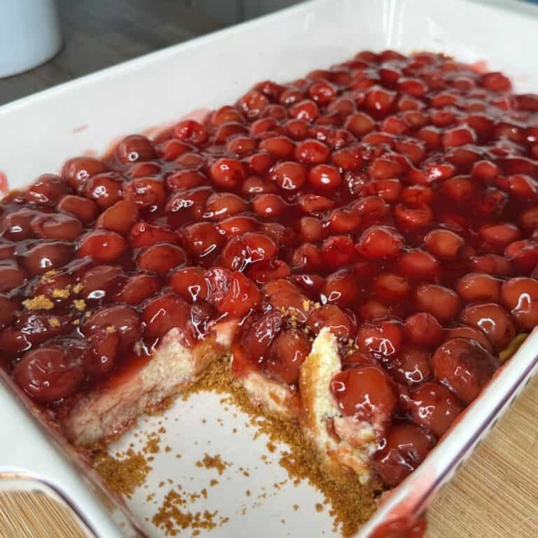 A rectangular baking dish filled with cherry dessert topped with glossy red cherries; a section has been cut out, revealing a crumbly base and creamy middle layer.