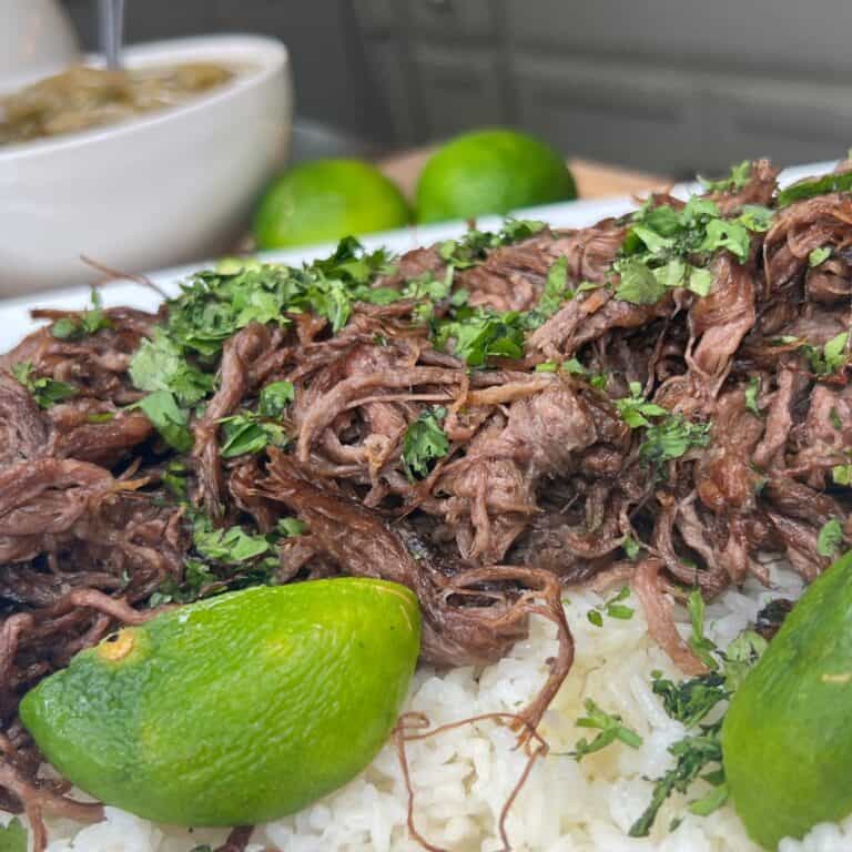 Shredded beef garnished with chopped cilantro served over white rice, with fresh lime wedges on the side. A bowl of green salsa is blurred in the background.